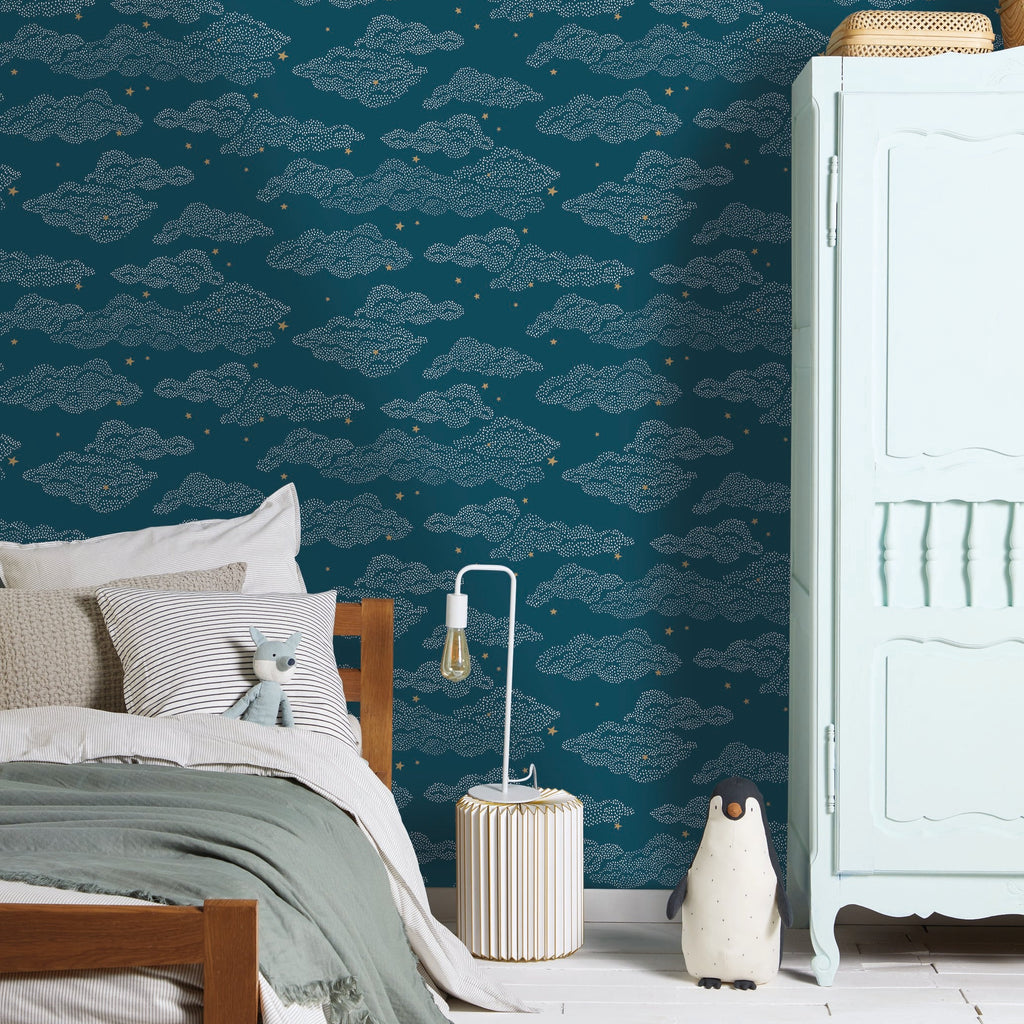 A styled bedroom corner featuring dark teal wallpaper with a repeating light blue cloud pattern and small yellow stars. A wooden bed with white and sage green bedding is on the left, a white cabinet on the right, and a striped rug on the floor.