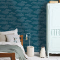 A styled bedroom corner featuring dark teal wallpaper with a repeating light blue cloud pattern and small yellow stars. A wooden bed with white and sage green bedding is on the left, a white cabinet on the right, and a striped rug on the floor.