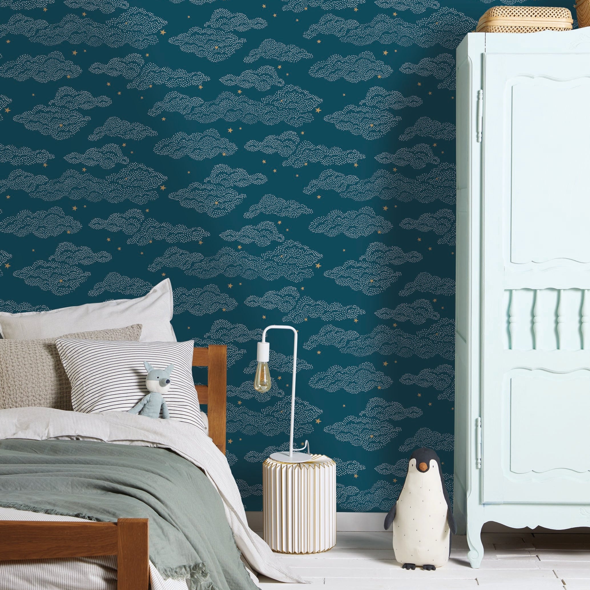 A styled bedroom corner featuring dark teal wallpaper with a repeating light blue cloud pattern and small yellow stars. A wooden bed with white and sage green bedding is on the left, a white cabinet on the right, and a striped rug on the floor.