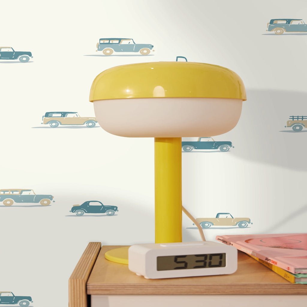 A retro mushroom-shaped table lamp with a yellow top and stem, and a cream-colored base, shown on a wooden bedside table next to a digital alarm clock and a pink book against a patterned wallpaper background.