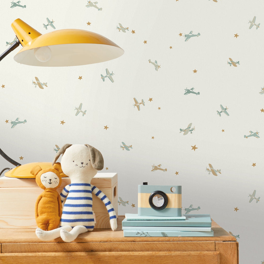 A styled children's room corner featuring a cream wallpaper with a pattern of small blue birds and gold stars, styled with a yellow desk lamp, a wooden chest of drawers, a plush dog toy, and a vintage camera.