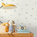 A styled children's room corner featuring a cream wallpaper with a pattern of small blue birds and gold stars, styled with a yellow desk lamp, a wooden chest of drawers, a plush dog toy, and a vintage camera.